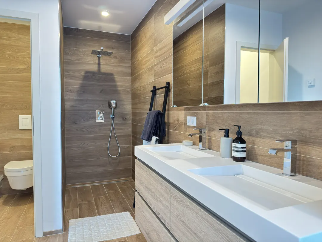 Salle de bains attenante avec douche, double vasque, miroir et WC, carrelage effet bois