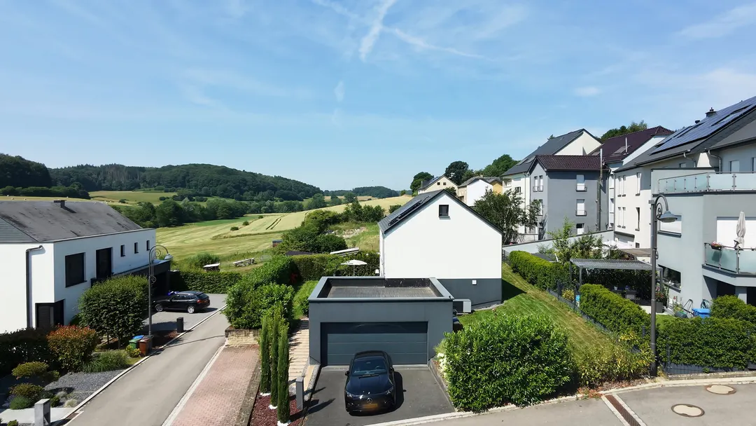 Vue aérienne de la maison blanche et de son garage, prairie et collines boisées derrière