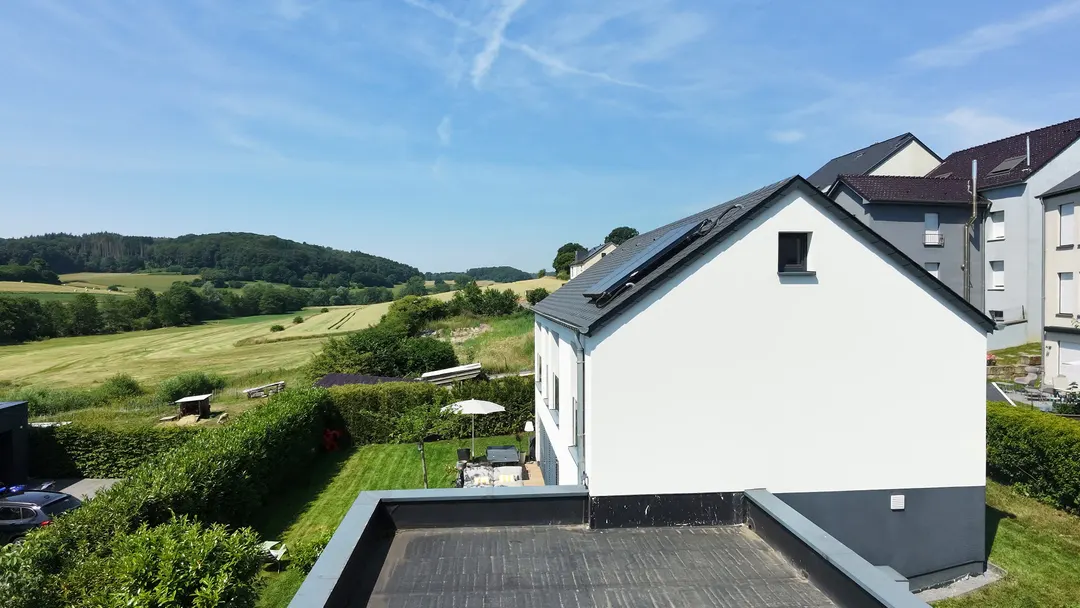 Pignon de la maison avec panneaux solaires, toit plat du garage au premier plan, vallée en fond