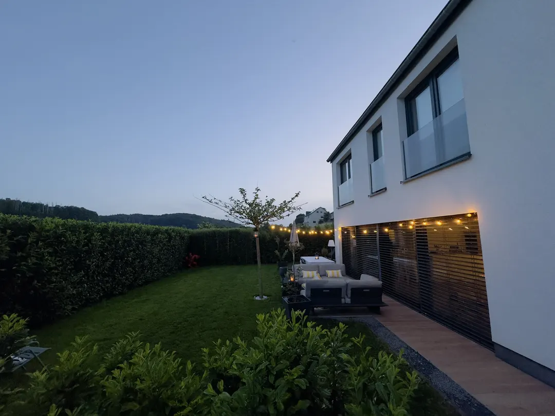 Jardin au crépuscule, terrasse éclairée, haie et collines boisées derrière