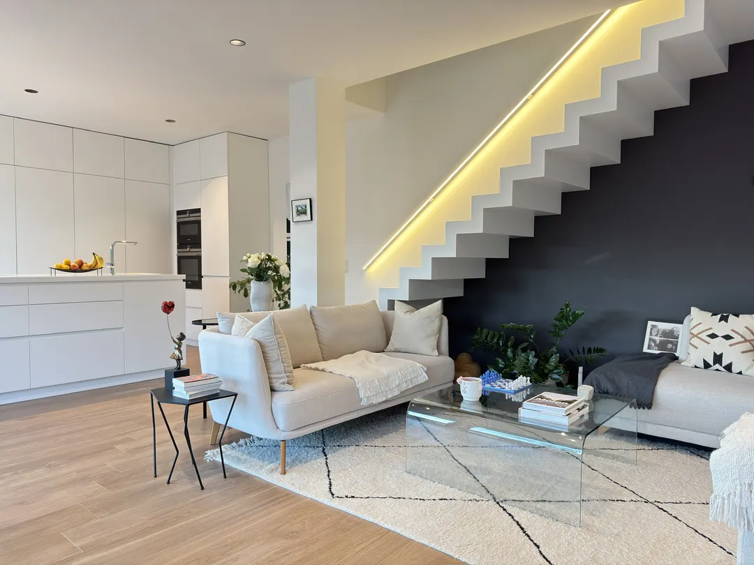 Pièce de vie ouverte : îlot de cuisine, mur de placards avec fours, canapé et escalier